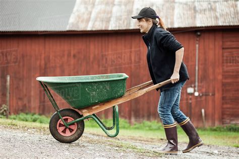 Side view of female farmer pushing wheelbarrow on rural road - Stock Photo - Dissolve