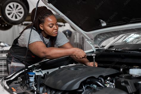 Black female mechanic working under the hood at repair garage. Portrait of confident focused ...