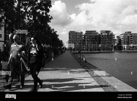 A grayscale of people walking by water at Grand Canal Dock with modern building in the ...