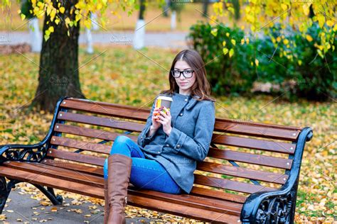 Beautiful young woman sitting on a bench drinking coffee or hot tea in the spring autumn coat ...