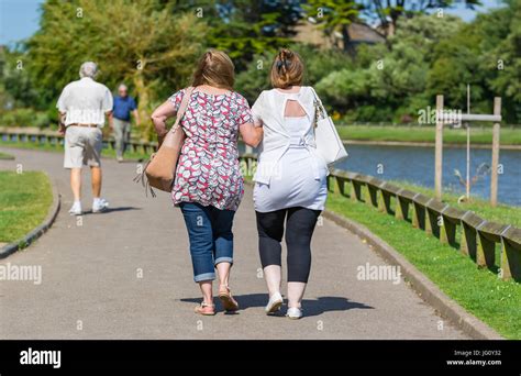 Female friends linking arms while walking through a park by a lake on a warm summer's day Stock ...