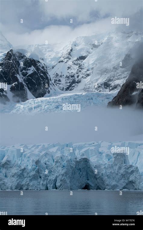 Fog cuts through the middle of a glacier crowned by mountains and clouds, Paradise Bay (Paradise ...