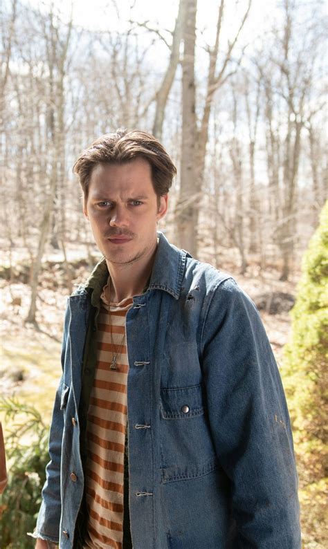 a man standing in the woods wearing a jean jacket and striped shirt with his hands in his pockets