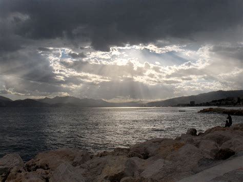 the sun shines through dark clouds over water and rocky shore with mountains in the distance
