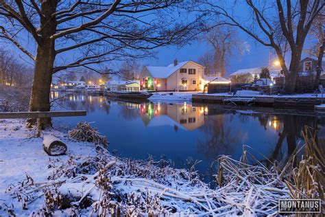 a small lake surrounded by snow covered trees and houses in the background at night with lights on