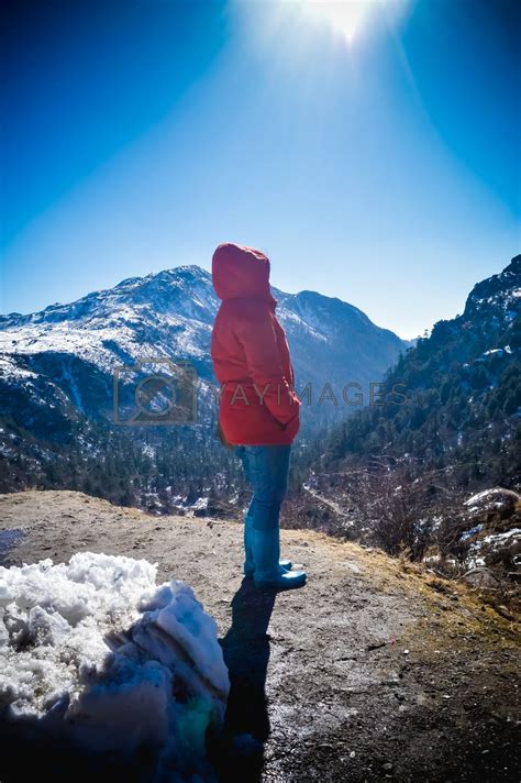 A woman in winter clothing standing on top of the rock of a snowcapped rocky mountain. Rear view ...