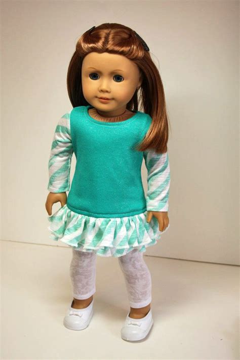 a doll with brown hair wearing a green shirt and white leggings, standing in front of a white ...