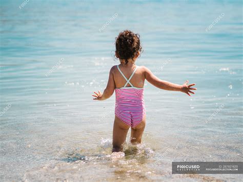 Back view of anonymous joyful cute girl playing in water of sea on background of calm seashore ...