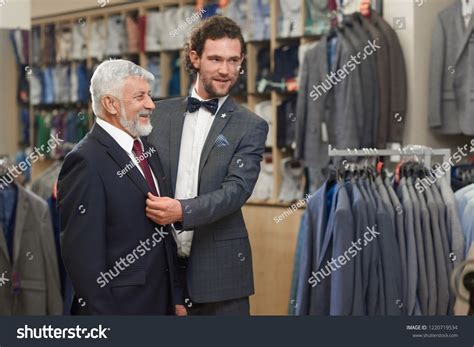 Attractive guy and older grey man in costumes buying clothes in male store. Salesman helping ...