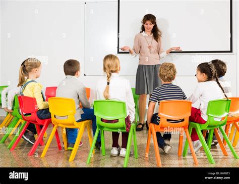 Portrait of young female teacher conducting lesson for small school children, listening ...