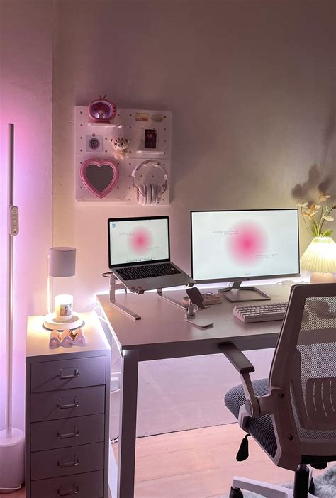 a desk with two computers on it in front of a pink light and some flowers