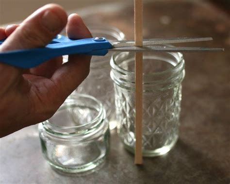 a person holding a pair of scissors in front of mason jars with sticks sticking out of them