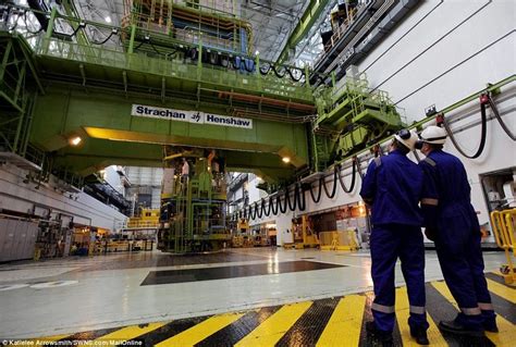 two men in blue work on the inside of a large factory with yellow and black stripes