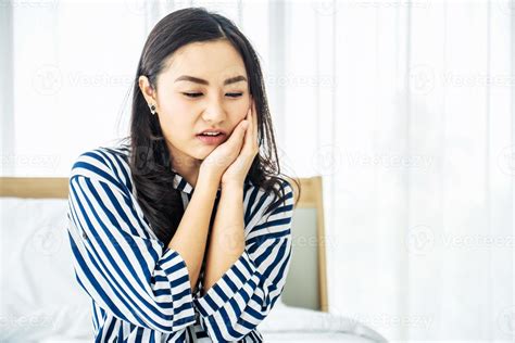 Young Asian woman touching cheek while suffering from toothache and discomfort on bed in white ...