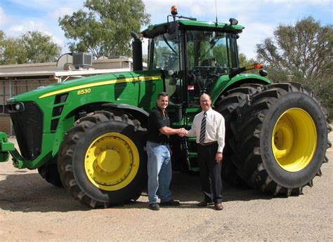 two men shaking hands in front of a large green tractor with yellow tires and rims