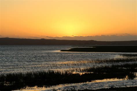 the sun is setting over the water and hills are in the distance with grass on either side