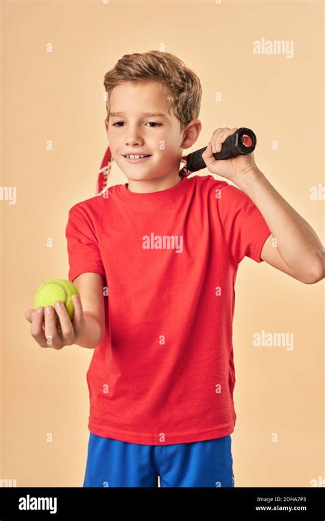 Cute male child tennis player holding yellow ball and smiling while standing against light ...
