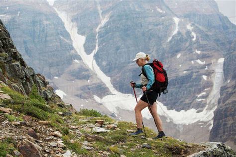 Female hiker with poles hiking up mountain path with mountain cliff with snow in background ...