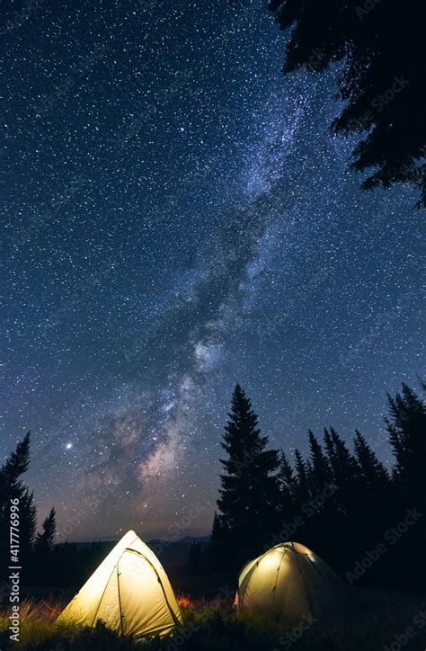 Vertical shot of tourist camping in forest, warm summer evening. Two illuminated tents under ...