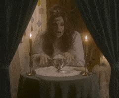 Video gif. Woman dressed as a fortune teller sits at a circular table with candles and tarot ...