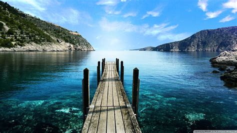 a wooden dock extending out into the ocean with mountains in the background and blue skies above