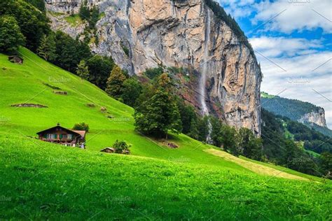 a green valley with a waterfall in the background and houses on the hill below it