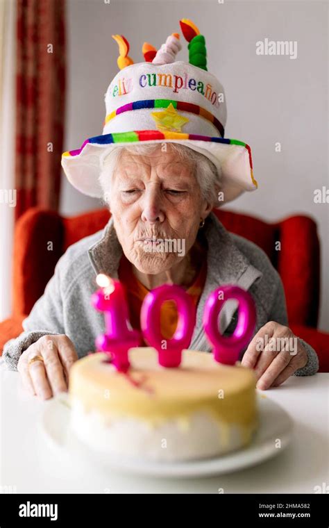 Elderly female in funny hat blowing burning candles with number 100 on birthday cake and ...