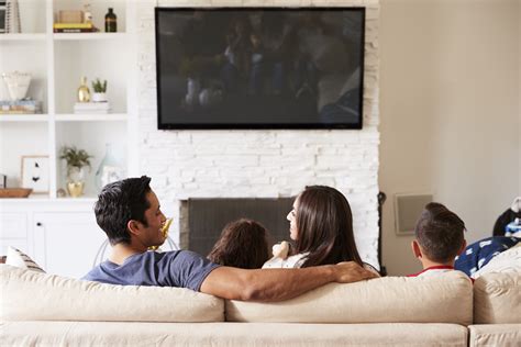 Back View Of Young Hispanic Family Of Four Sitting On The Sofa Watching TV, Mum Looking At Dad ...