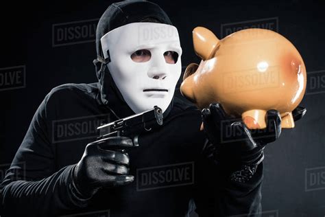 Robber in balaclava and mask aiming at piggy bank - Stock Photo - Dissolve