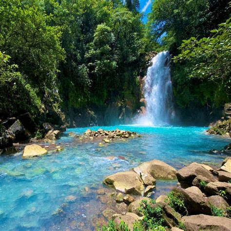 a waterfall in the middle of a river surrounded by rocks and greenery with blue water