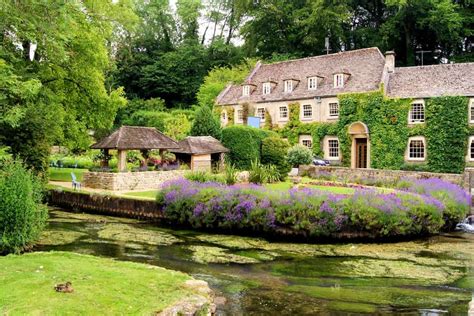 an old house is surrounded by flowers and greenery in the foreground with a stream running ...