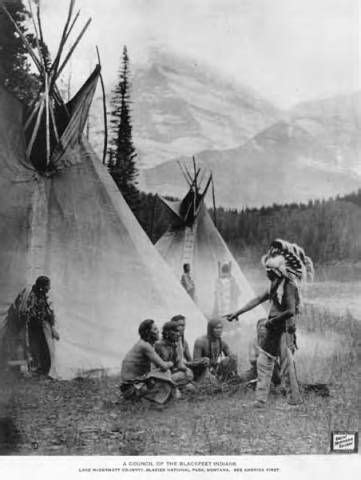 Group of seated Piegan Blackfeet men and a standing woman listening to a man in ceremonial ...