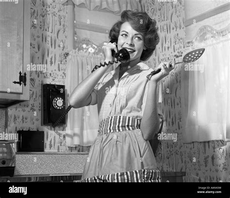 1950s 1960s SMILING WOMAN TALKING WALL TELEPHONE HOLDING SPOON WEARING APRON STANDING IN KITCHEN ...