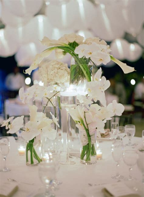 white flowers and candles are arranged in clear vases on a round table with glassware