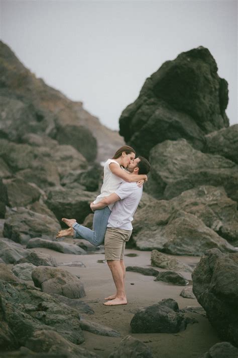 A couple on an engagement photo shoot at Muir Beach kissing each other on the beach in front of ...