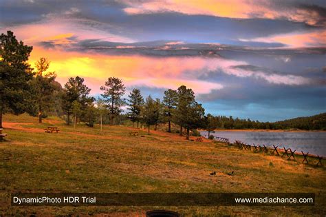 a beautiful sunset over a lake with benches on the grass and trees in ...