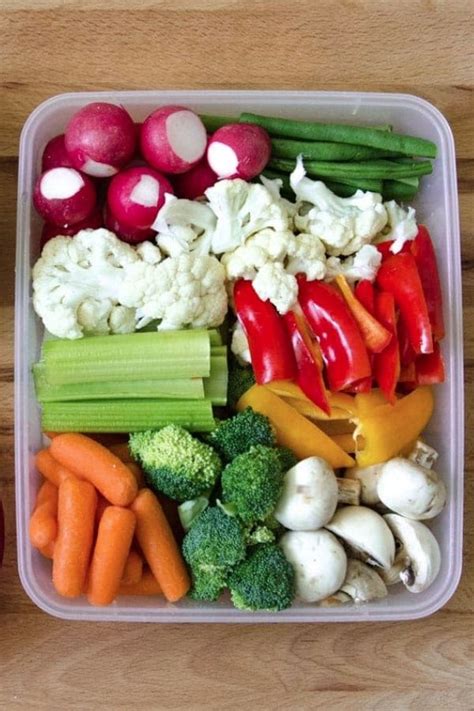 a plastic container filled with veggies on top of a wooden table next to an apple