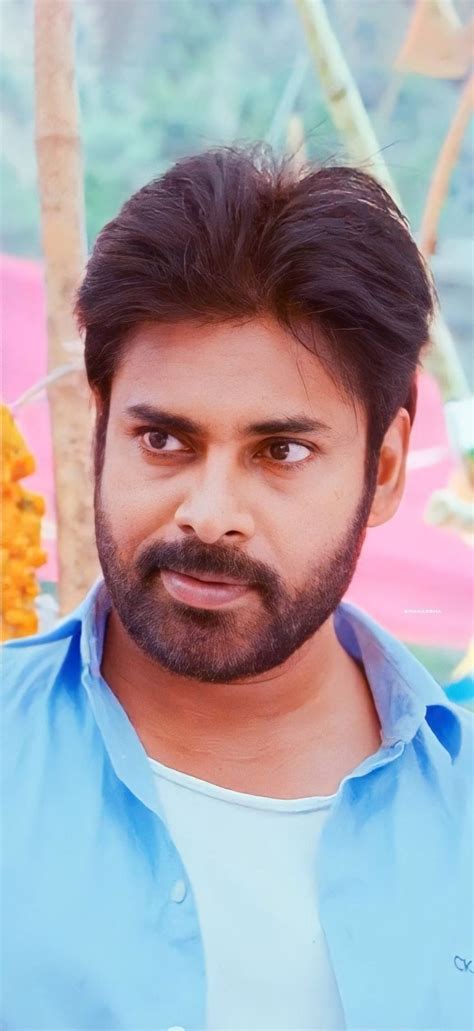 a man with a beard and blue shirt looks at the camera while standing in front of flowers