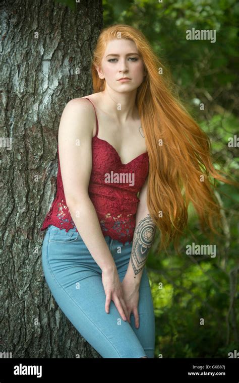 Beautiful red head woman in skinny jeans and red top, leaning nearly full length against a tree ...