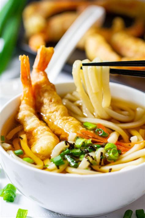 Close up of a bowl of udon soup with shrimp tempura on top , chopsticks lifting the noodles, and ...