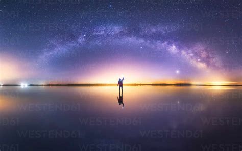 Silhouette of anonymous man standing on reflection surface of water and reaching out to starry ...