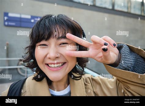 Positive korean girl in headphones, drinks coffee to go, shows peace, v-sign, stands on street ...