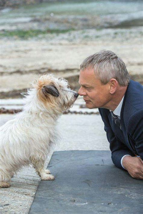 a man in suit and tie kissing a small white dog on the ground next to water