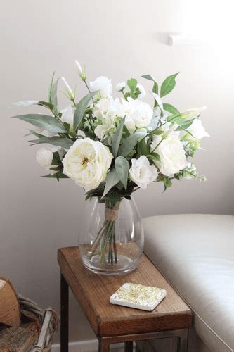 a glass vase with white flowers on a small table in front of a couch and chair