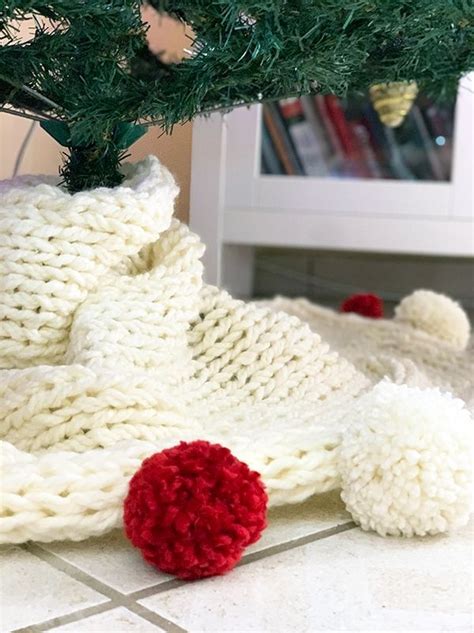 a knitted christmas tree skirt with pom - poms sits on the floor