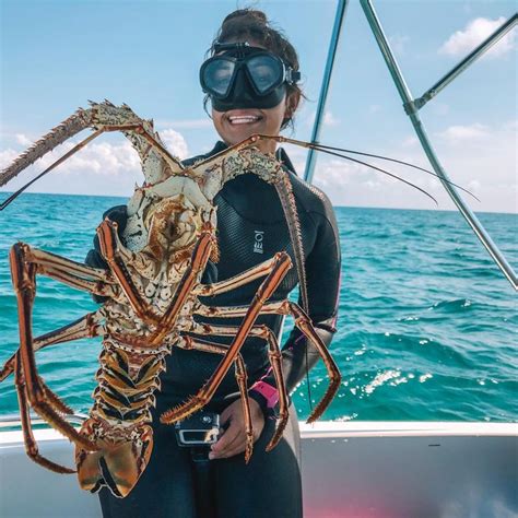 a woman in a wetsuit holding up a large lobster on a boat with the ocean behind her