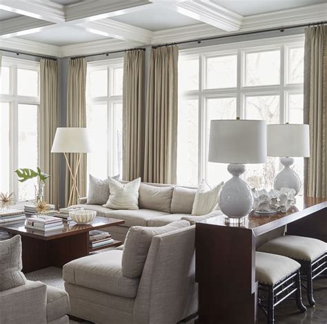 neutral living room; white lamps; large windows; curtains; grey couches; beige couches; exposed ...