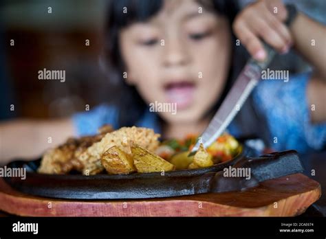 Little girl holding knife and fork cutting grilled Chicken steak on stoned plate in restaurant ...