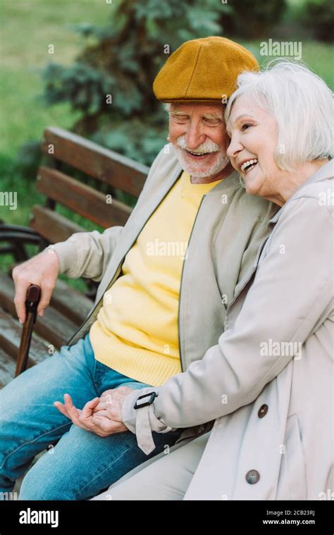 Smiling elderly couple holding hands while sitting on bench in park ...