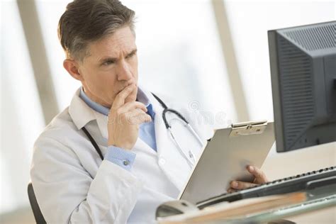 Serious Doctor Looking At Computer While Holding Clipboard At De Stock Photo - Image of mature ...